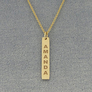 A gold bar necklace with the name 'AMANDA' vertically engraved, displayed on a textured gray background.