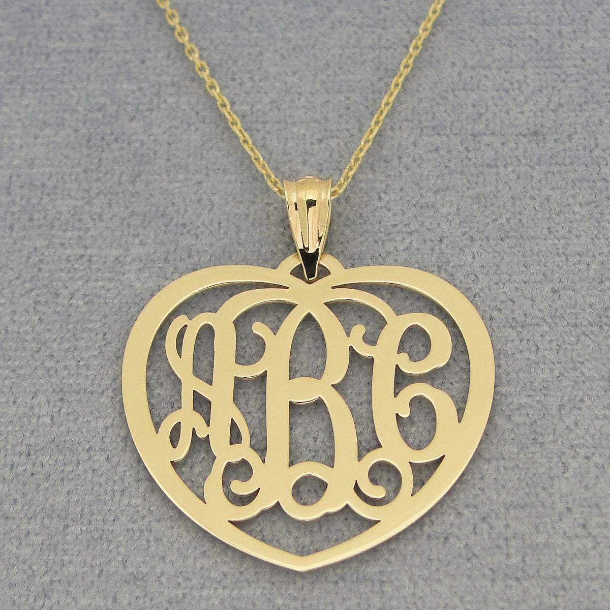 Close-up of custom solid gold heart monogram pendant with three initials on a gold chain and gray background