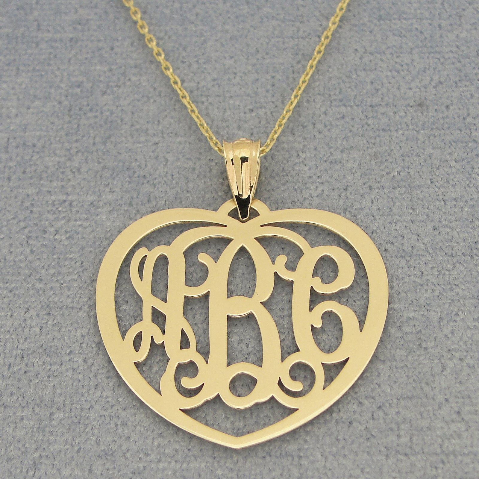 Close-up of custom solid gold heart monogram pendant with three initials on a gold chain and gray background
