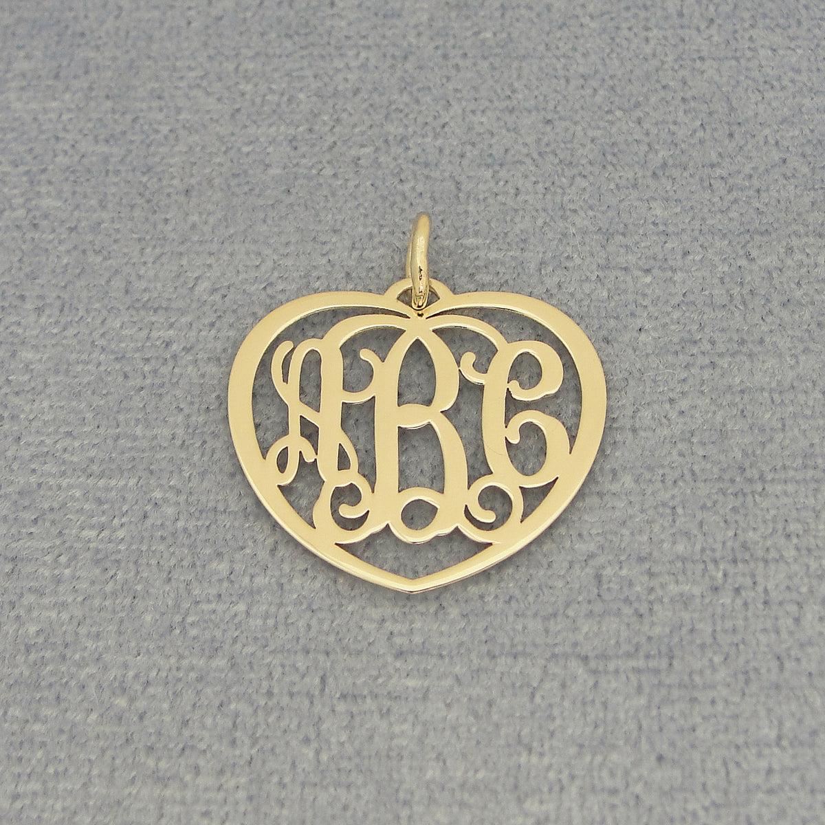Close-up of a small gold heart monogram pendant with three custom initials on a gray background.