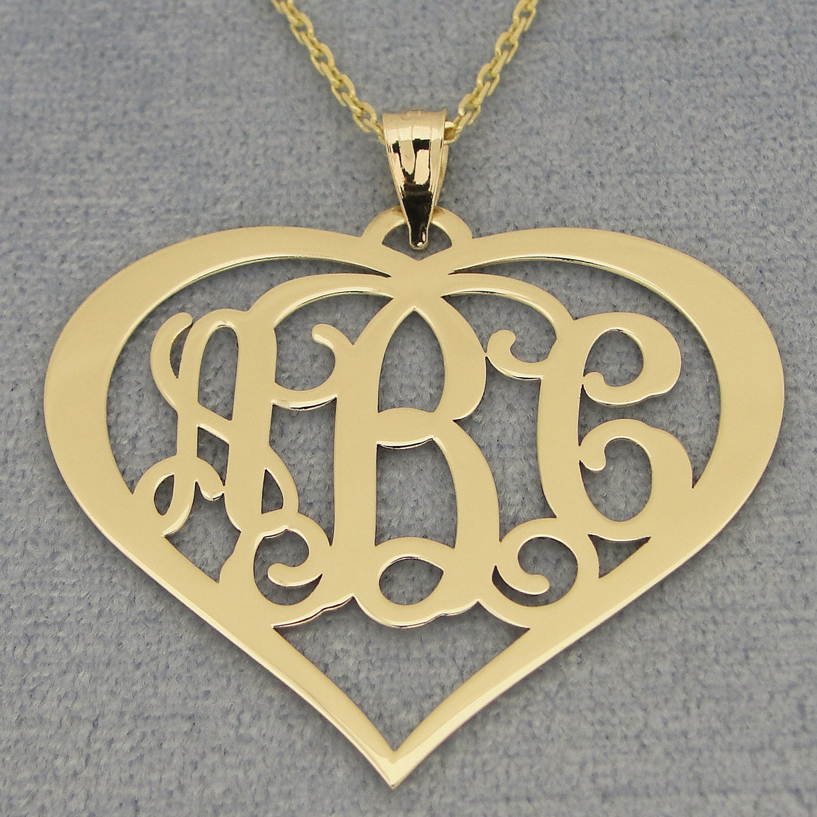 Solid gold 3-initial heart monogram necklace with a chain on a gray background.