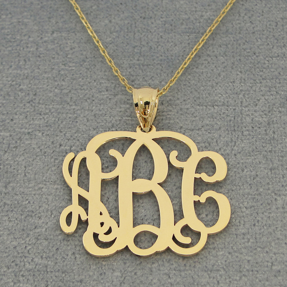 Solid yellow gold three-initial monogram pendant on a fine chain against a gray jewelry background.