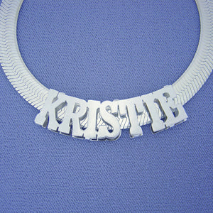 Personalized 3D sterling silver block nameplate necklace spelling "KRISTIE" on a thick silver herringbone chain, displayed against a blue background.