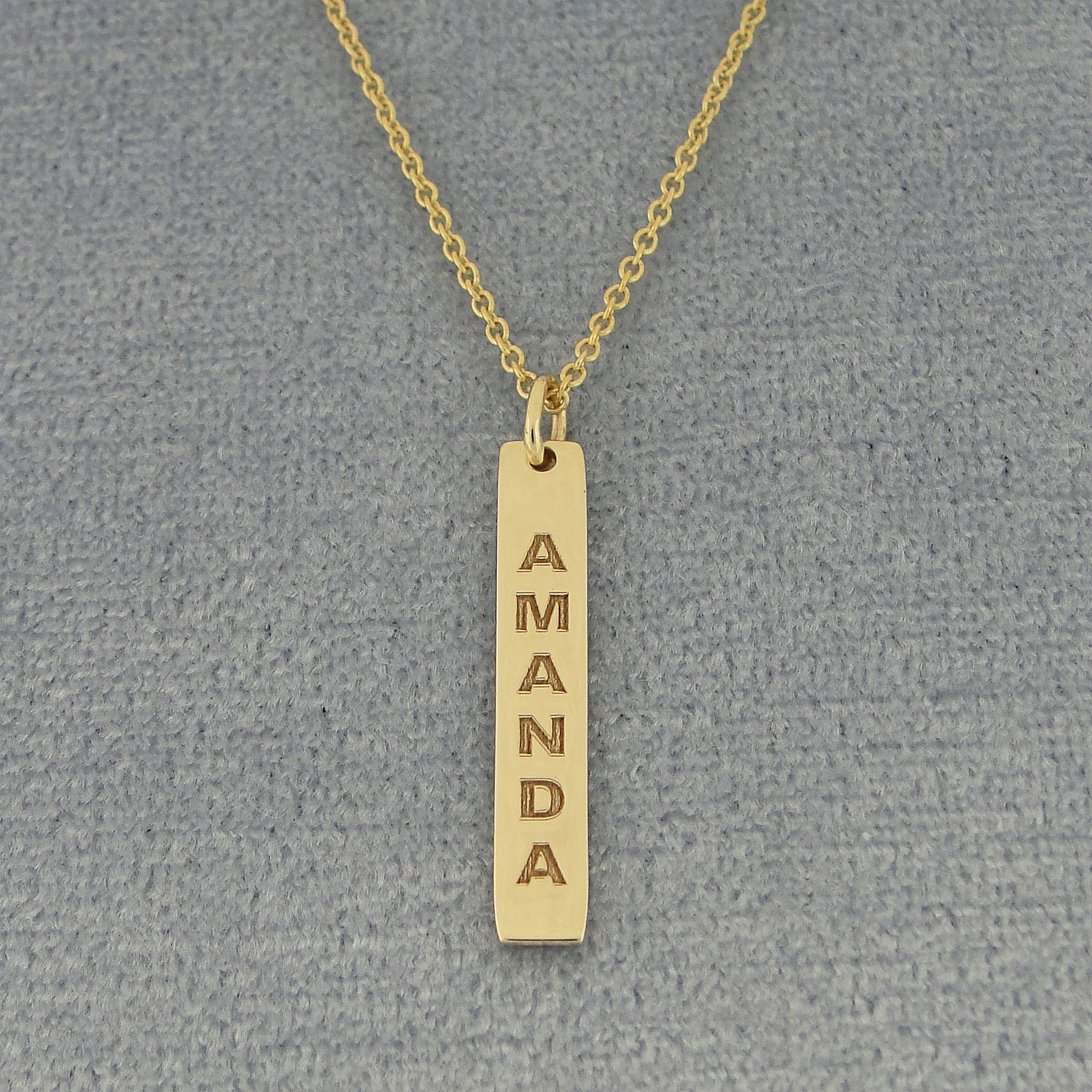 A gold bar necklace with the name 'AMANDA' vertically engraved, displayed on a textured gray background.