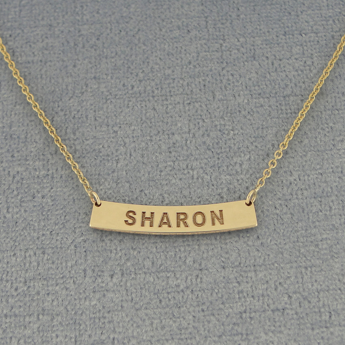 A gold bar necklace with the name 'SHARON' engraved on a curved horizontal bar.