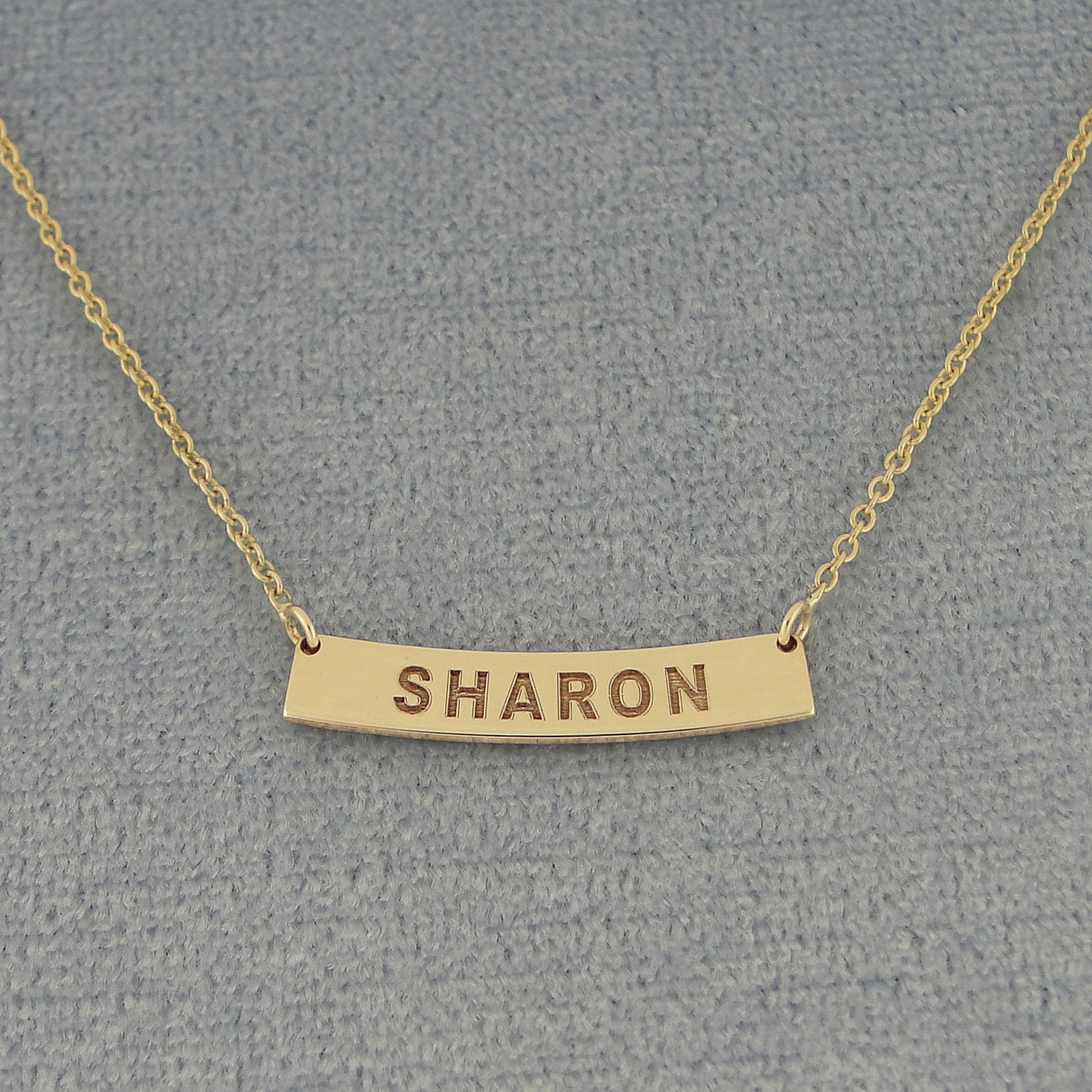 A gold bar necklace with the name 'SHARON' engraved on a curved horizontal bar.