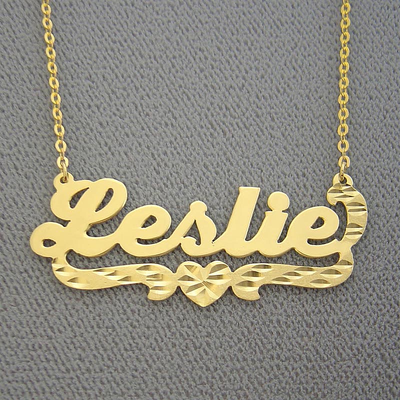 Custom solid gold nameplate necklace with "Leslie" written in script font featuring diamond-cut detailing and a heart design on a gold Rolo chain.