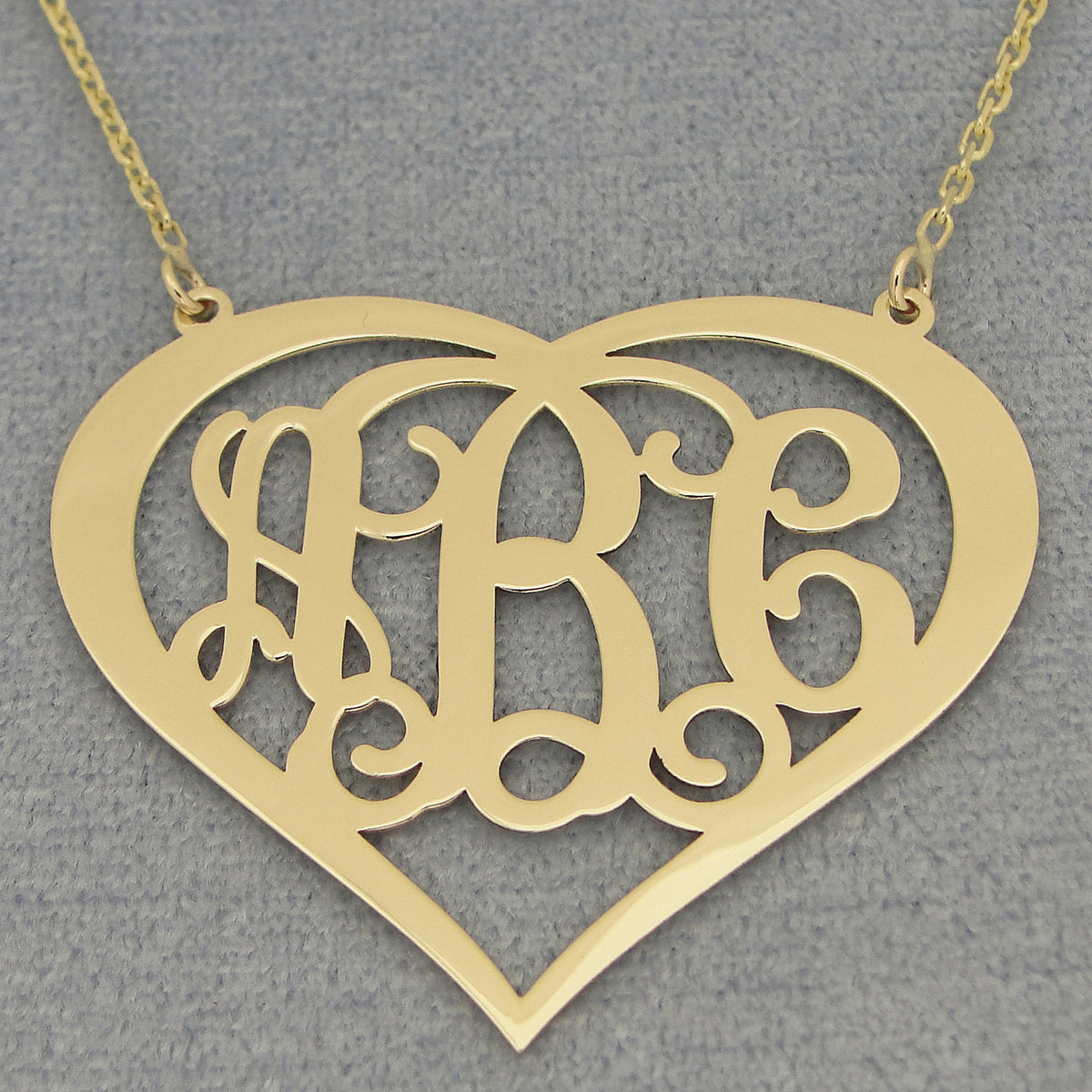 Close-up of a custom yellow solid gold three-initial heart monogram pendant on a gray background