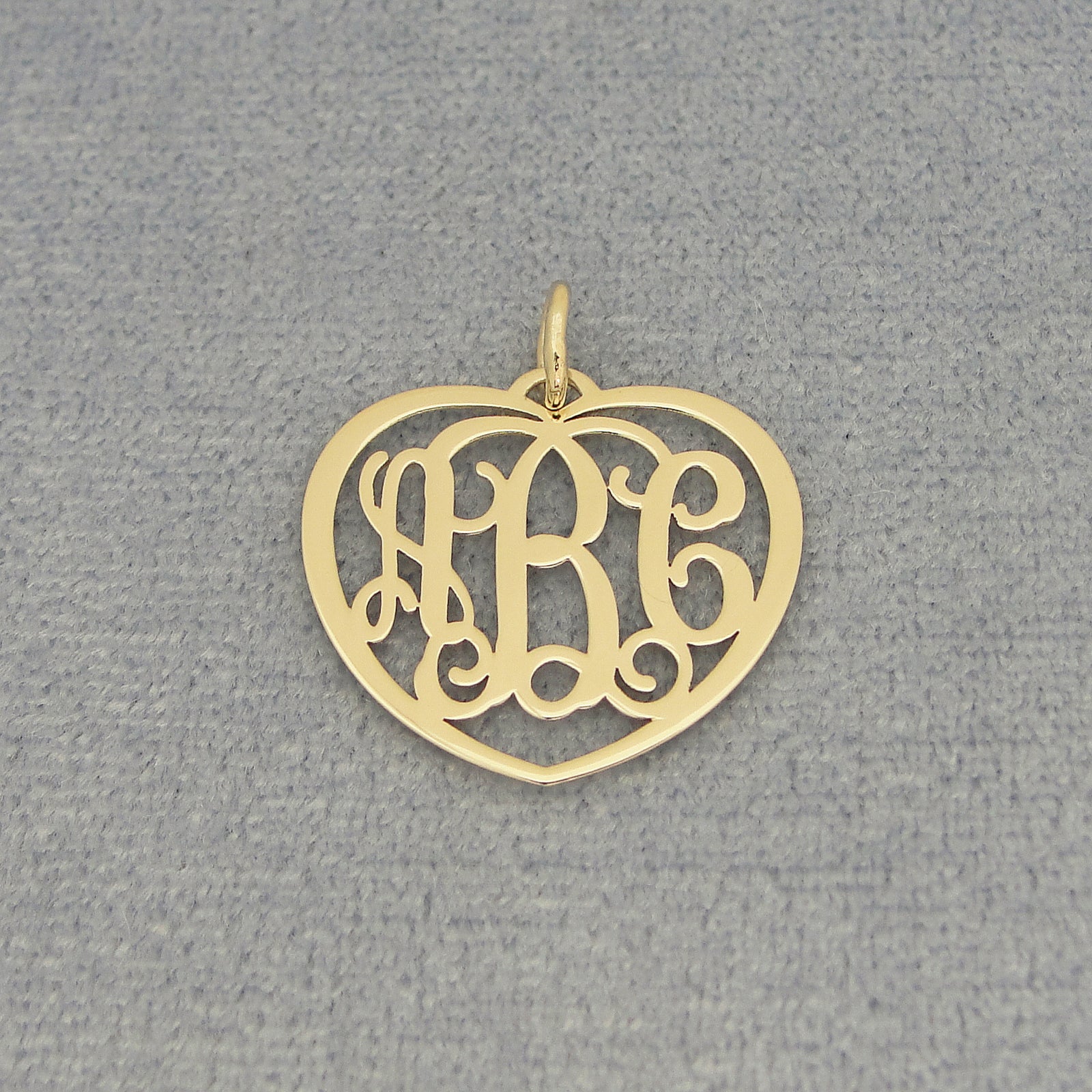 Close-up of a small gold heart monogram pendant with three custom initials on a gray background.