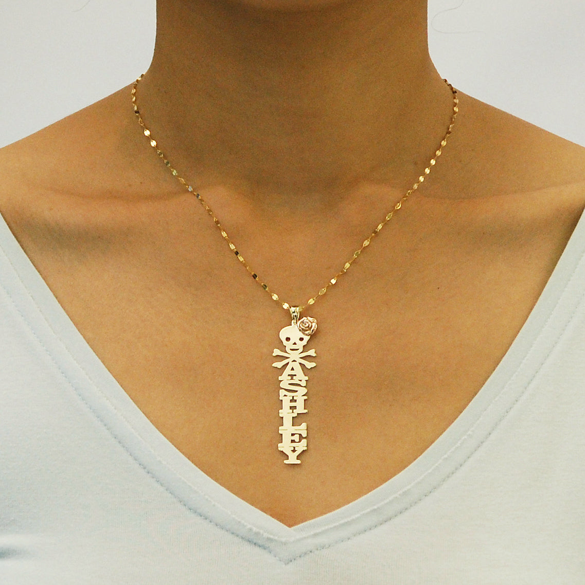 Close-up of an Ashley gold vertical name pendant with skull, crossbones, and rose detail, worn on a chain above a white V-neck top.