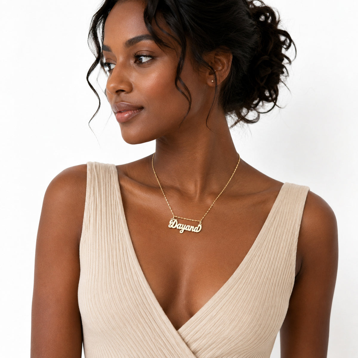 A woman wearing a delicate gold nameplate necklace that reads 'Dayana' on a fine chain, paired with a beige ribbed wrap top against a clean white background.